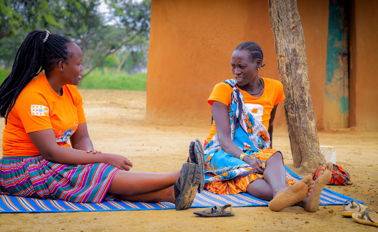 A woman wearing an orange t-shirt with a UNFPA logo speaks to Ms. Telo, who is also wearing orange. They are seated on a striped blanket in a courtyard.